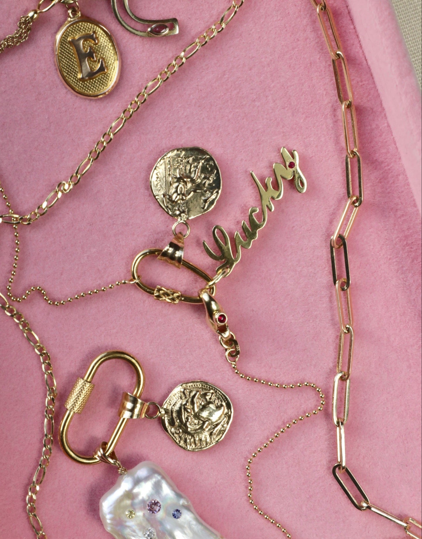 A variety of handcrafted charms and gold plated pendants, including a personalized pendant with the initial 'E' in gold, a small pearl gemstone pendant, and other decorative elements laid out on a pink surface.
